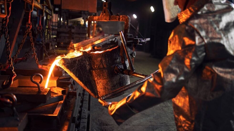 Molten metal pouring into a mold in a steel foundry, illuminated by orange glow and industrial equipment nearby.