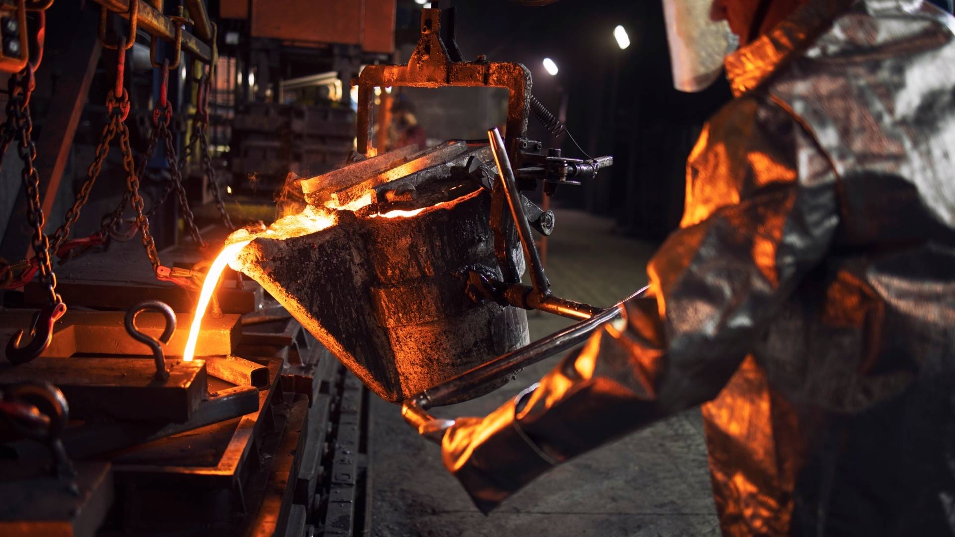 Molten metal pouring into a mold in a steel foundry, illuminated by orange glow and industrial equipment nearby.