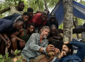 Group of men gathered outdoors around a bearded man with a tablet, under a blue tarp tent in a wooded campsite.