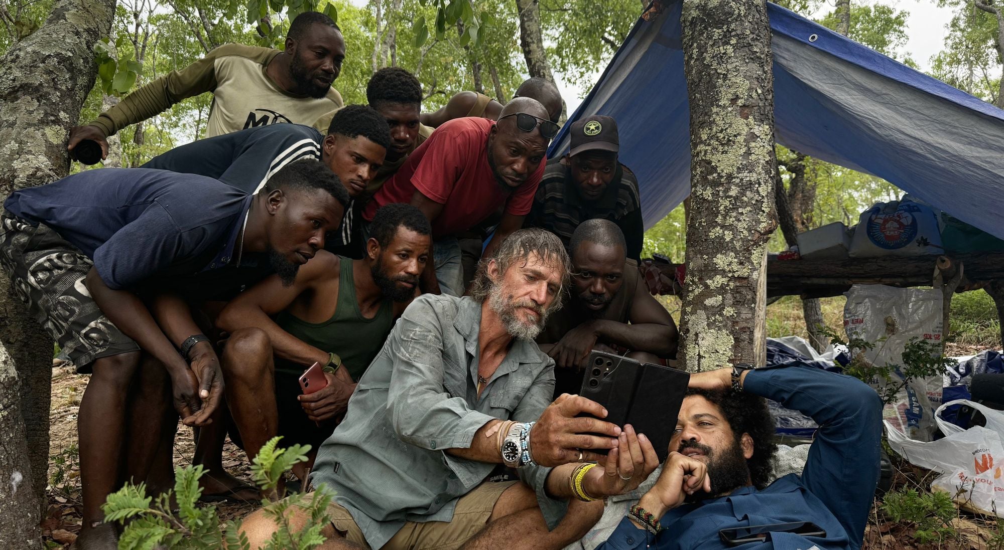 Group of men gathered outdoors around a bearded man with a tablet, under a blue tarp tent in a wooded campsite.