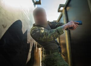 Tactical scene of a soldier in camouflage aiming a pistol through a narrow doorway, face blurred for privacy.