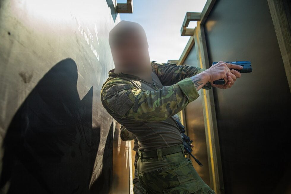 Tactical scene of a soldier in camouflage aiming a pistol through a narrow doorway, face blurred for privacy.