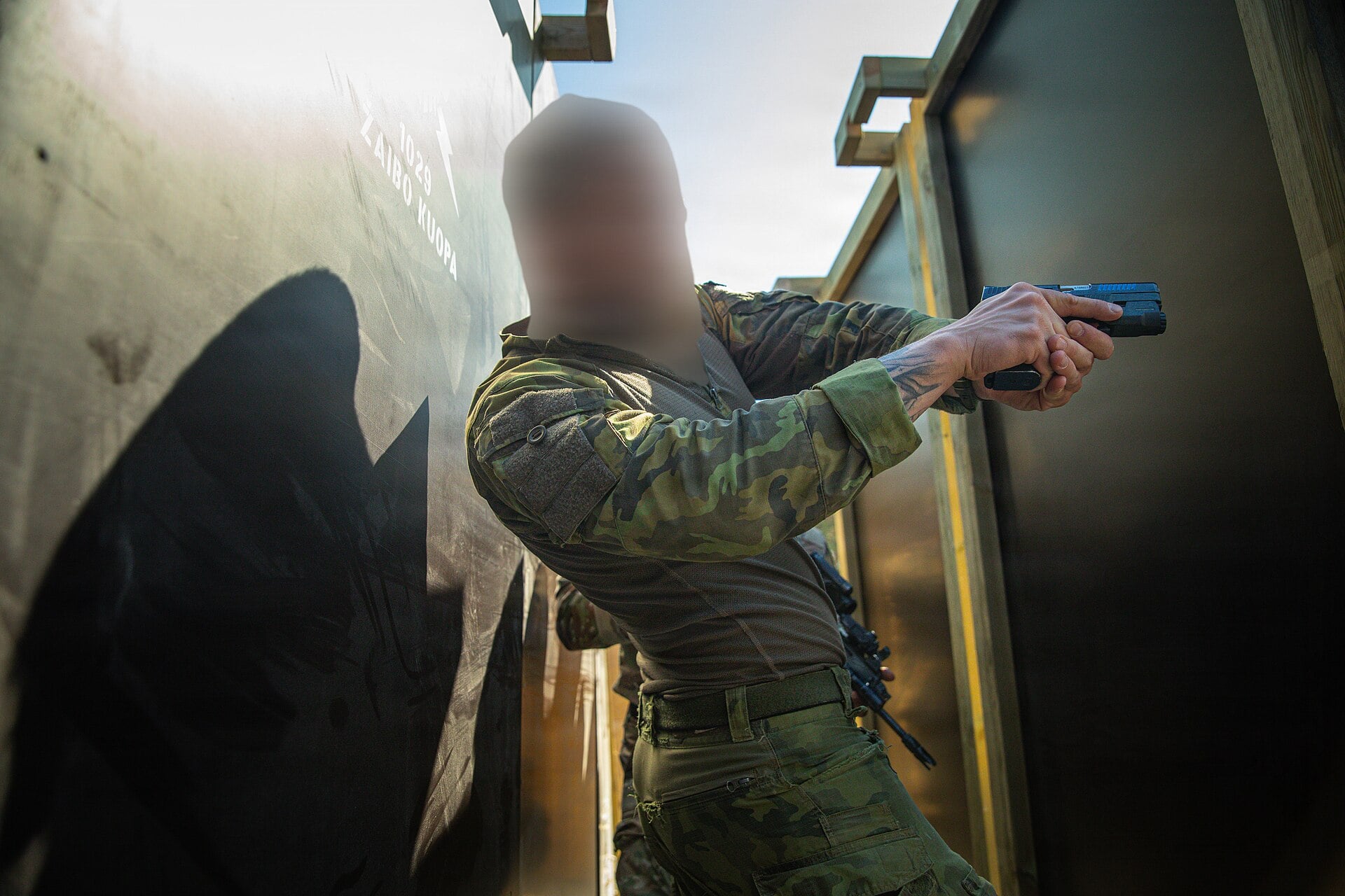 Tactical scene of a soldier in camouflage aiming a pistol through a narrow doorway, face blurred for privacy.