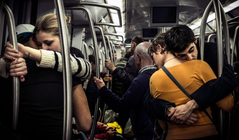 A couple shares a tight hug in a crowded subway car, standing beside metal poles and holding on for support.