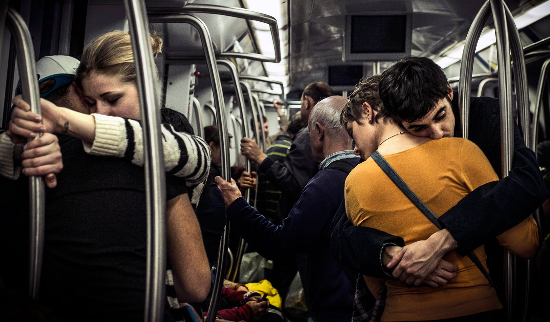 A couple shares a tight hug in a crowded subway car, standing beside metal poles and holding on for support.