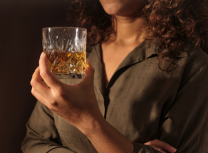 Person with curly hair in a dark green shirt holds a crystal tumbler filled with whiskey against a dark background.