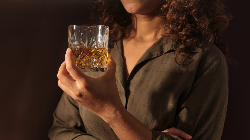 Person with curly hair in a dark green shirt holds a crystal tumbler filled with whiskey against a dark background.