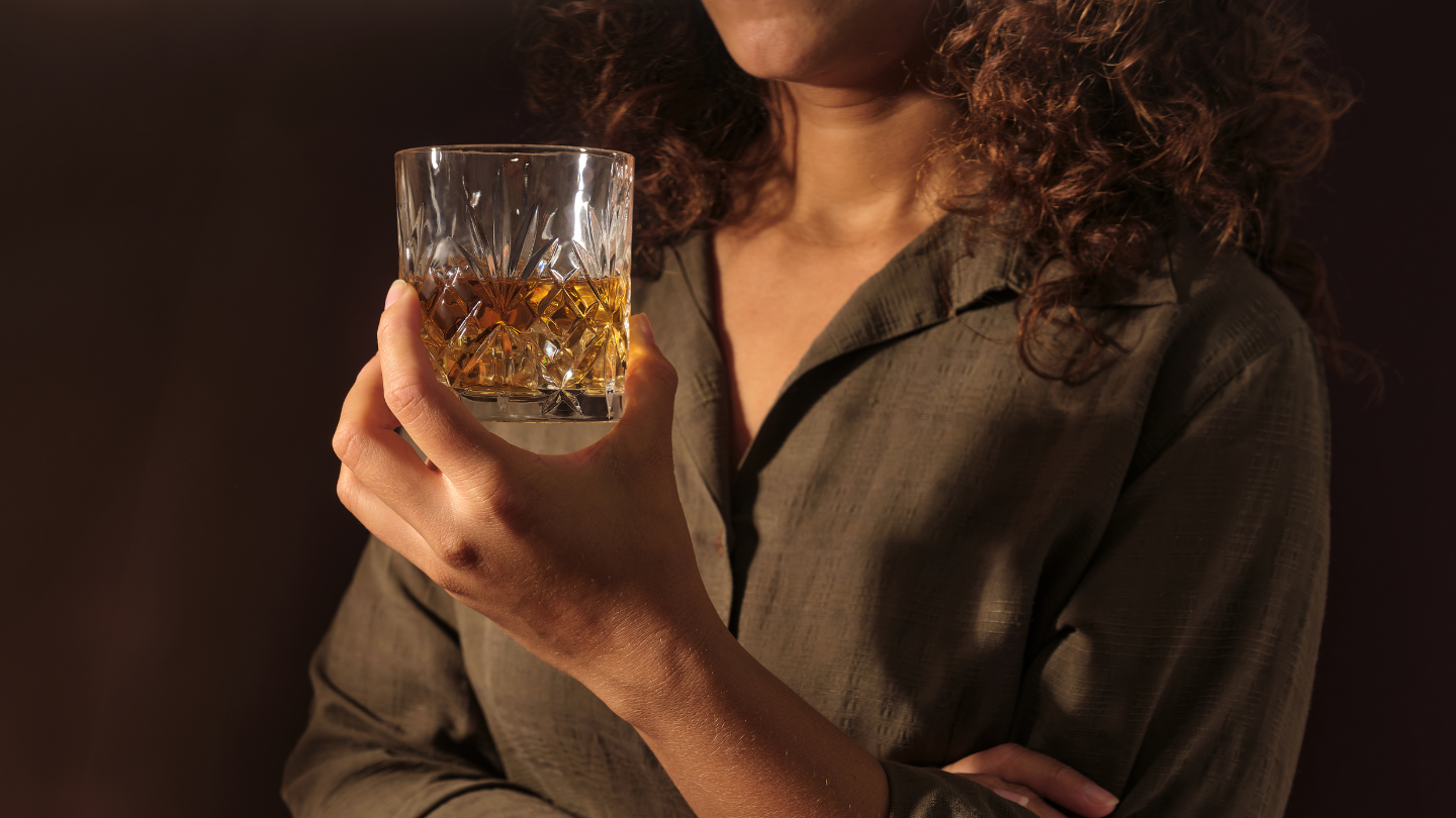 Person with curly hair in a dark green shirt holds a crystal tumbler filled with whiskey against a dark background.