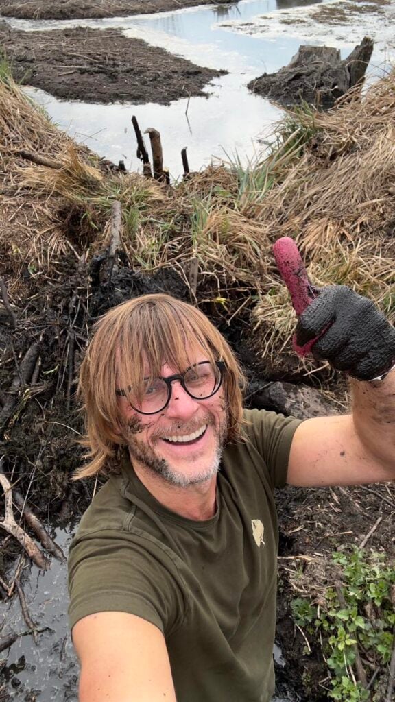 Smiling man with glasses and a muddy face gives a thumbs-up while taking a selfie near a muddy shoreline.