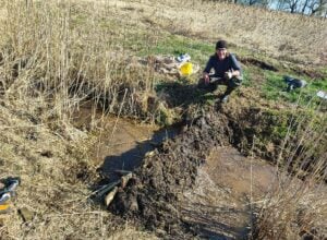 Man wearing a beanie and gloves crouches in a muddy marsh among tall dried reeds with gear nearby.] , but one should be careful with extra punctuation
