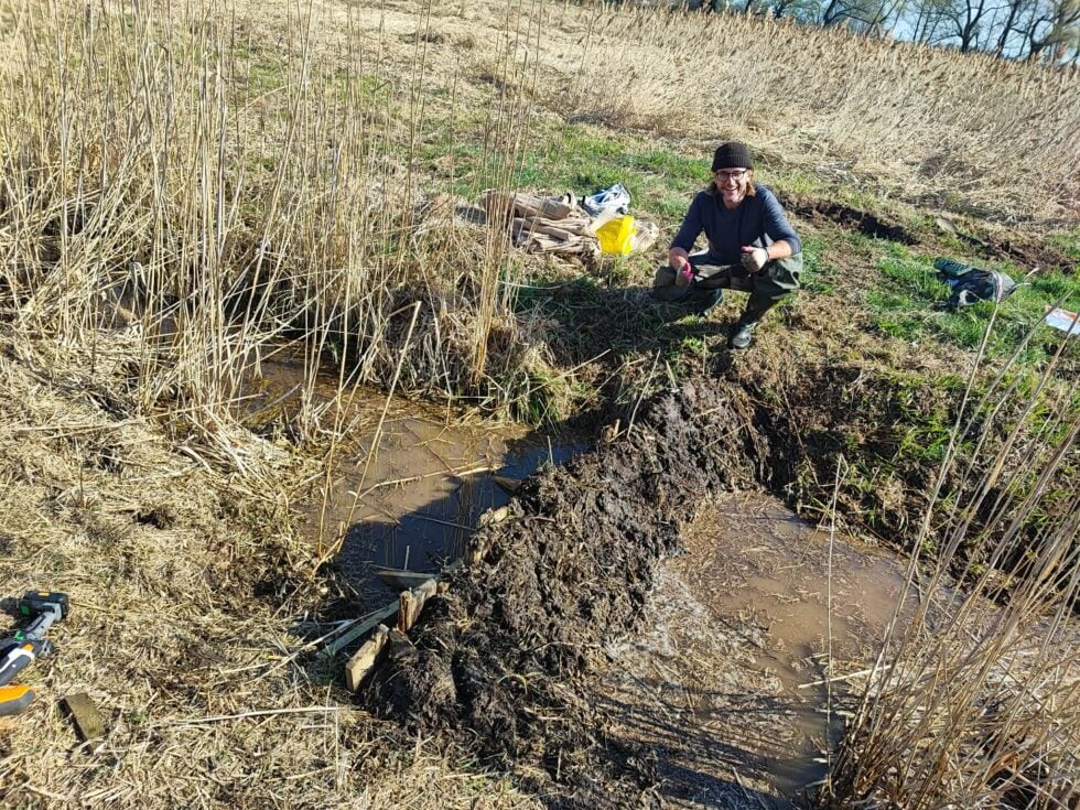 Man wearing a beanie and gloves crouches in a muddy marsh among tall dried reeds with gear nearby.] , but one should be careful with extra punctuation