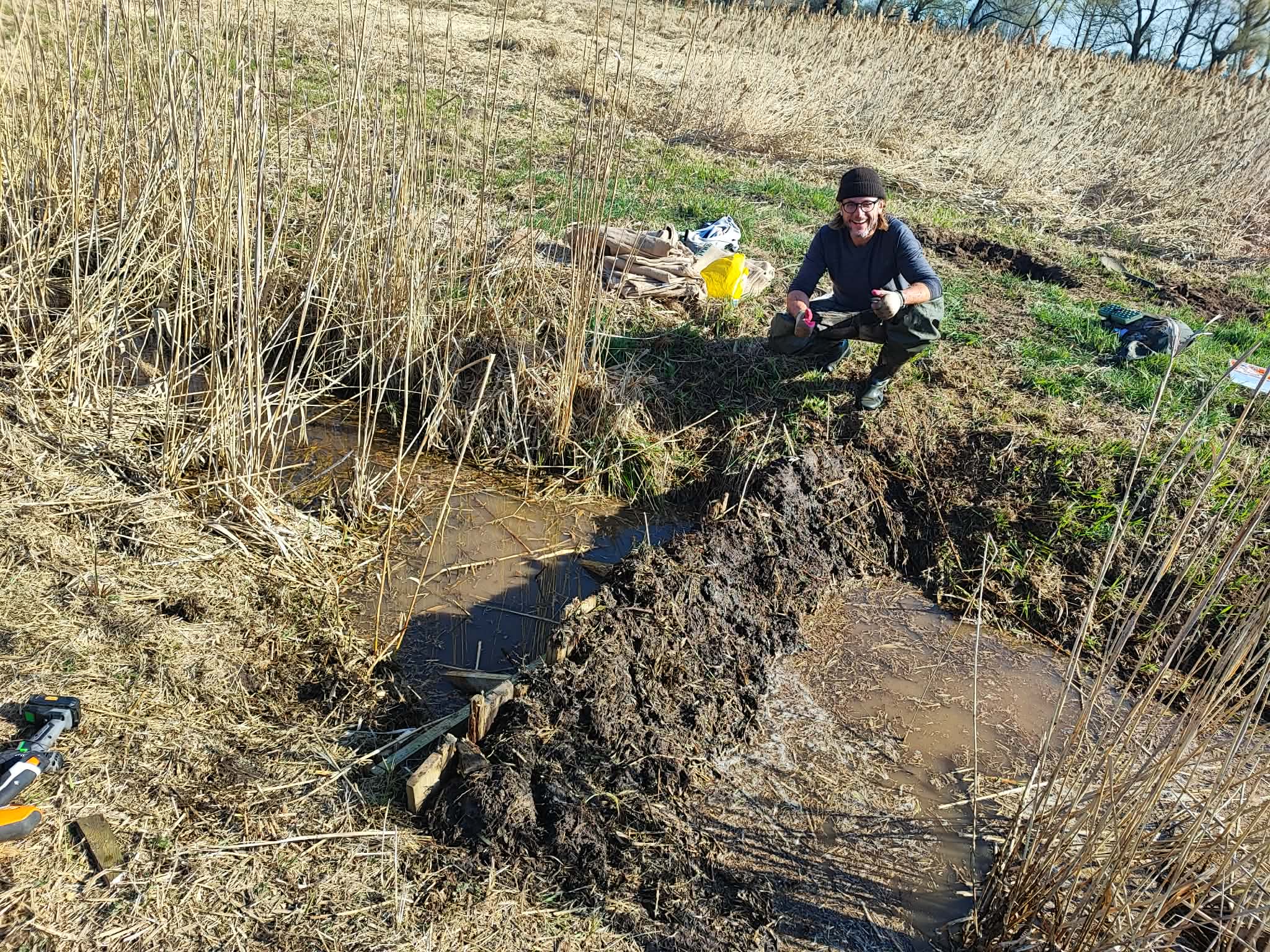Man wearing a beanie and gloves crouches in a muddy marsh among tall dried reeds with gear nearby.] , but one should be careful with extra punctuation