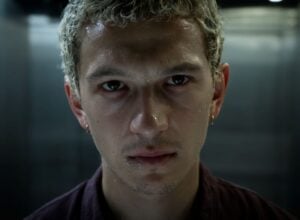 Close-up portrait of a young man with wet skin and intense gaze in a dim, metallic hallway backdrop.