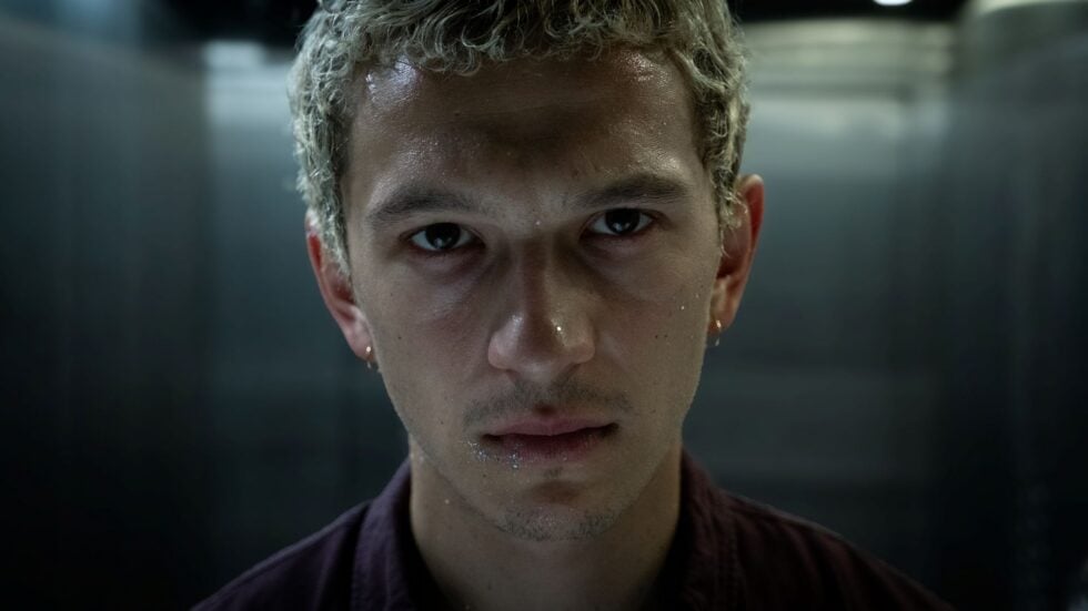 Close-up portrait of a young man with wet skin and intense gaze in a dim, metallic hallway backdrop.