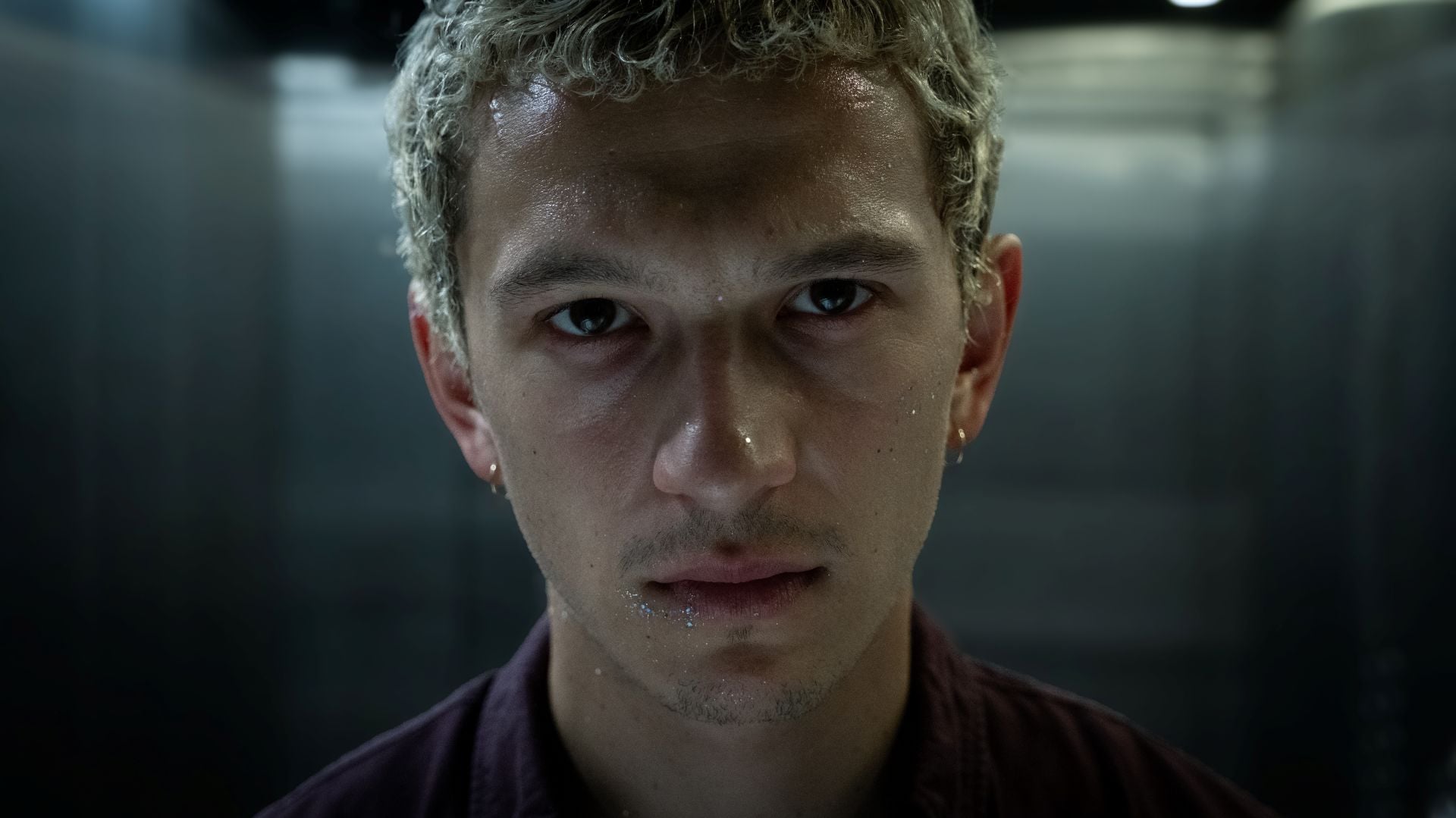 Close-up portrait of a young man with wet skin and intense gaze in a dim, metallic hallway backdrop.