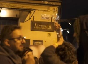Nighttime street scene outside a bar with a wooden 'ALCHEMIA' sign; a crowd raises glasses in a toast.