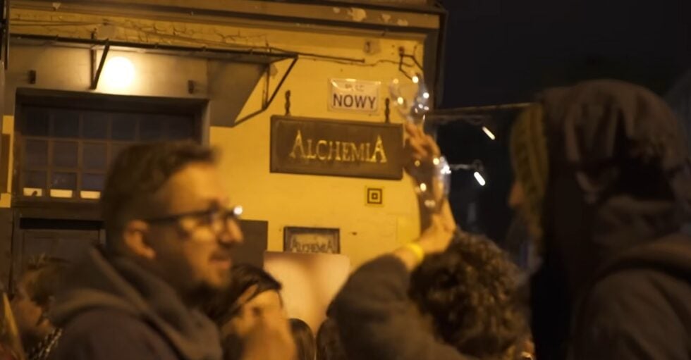 Nighttime street scene outside a bar with a wooden 'ALCHEMIA' sign; a crowd raises glasses in a toast.