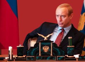 Man in a dark suit seated at an ornate desk with a gold eagle ornament, red backdrop, and flag banners in the background (formal office setting).