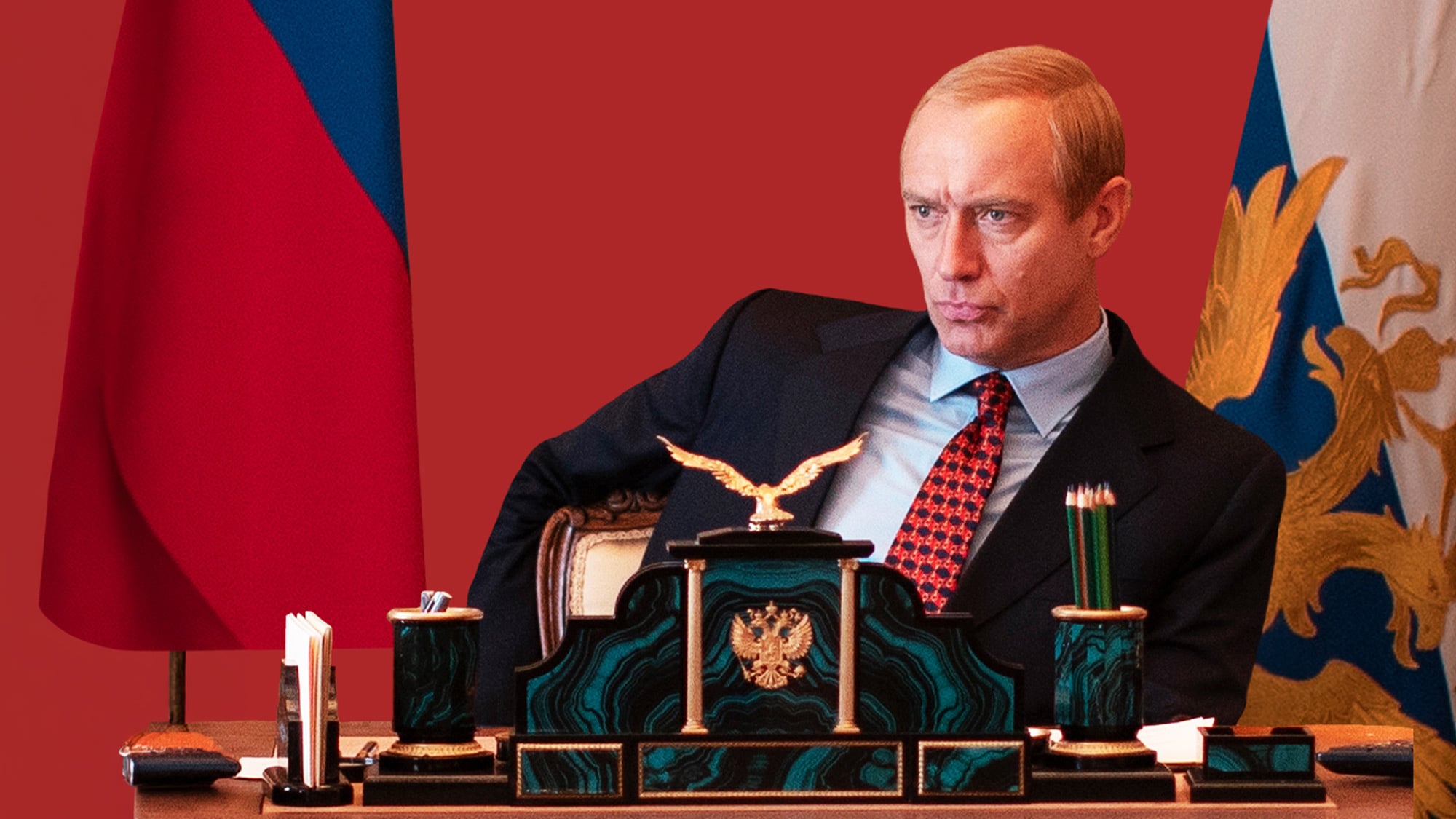 Man in a dark suit seated at an ornate desk with a gold eagle ornament, red backdrop, and flag banners in the background (formal office setting).