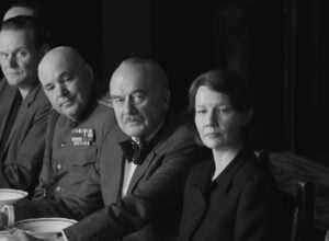 Black-and-white scene of people at a formal dinner table; a man in a decorated military uniform sits center, others beside him.