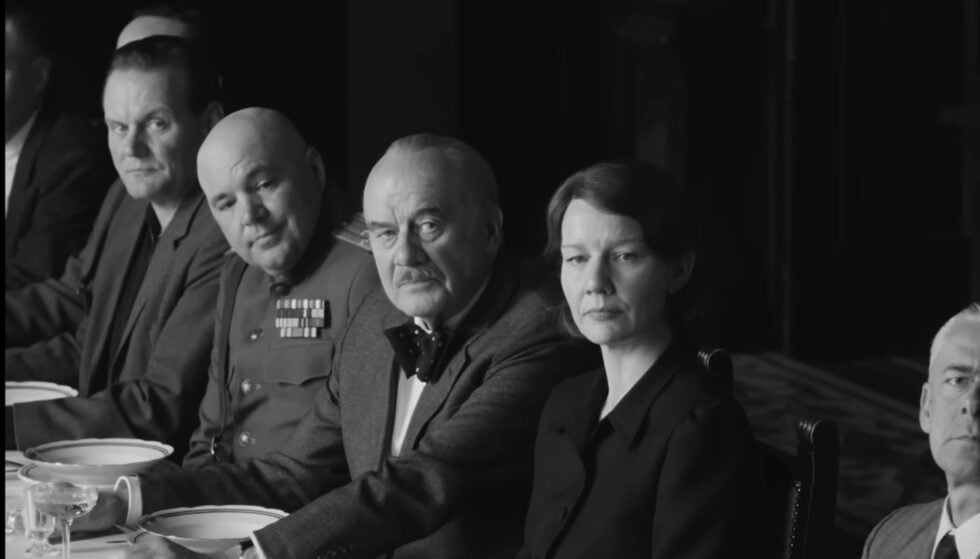 Black-and-white scene of people at a formal dinner table; a man in a decorated military uniform sits center, others beside him.