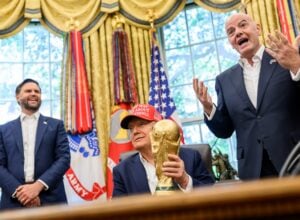 Three men in dark suits celebrate in a room with gold drapes and flags; one holds a large gold trophy while another gestures enthusiastically.
