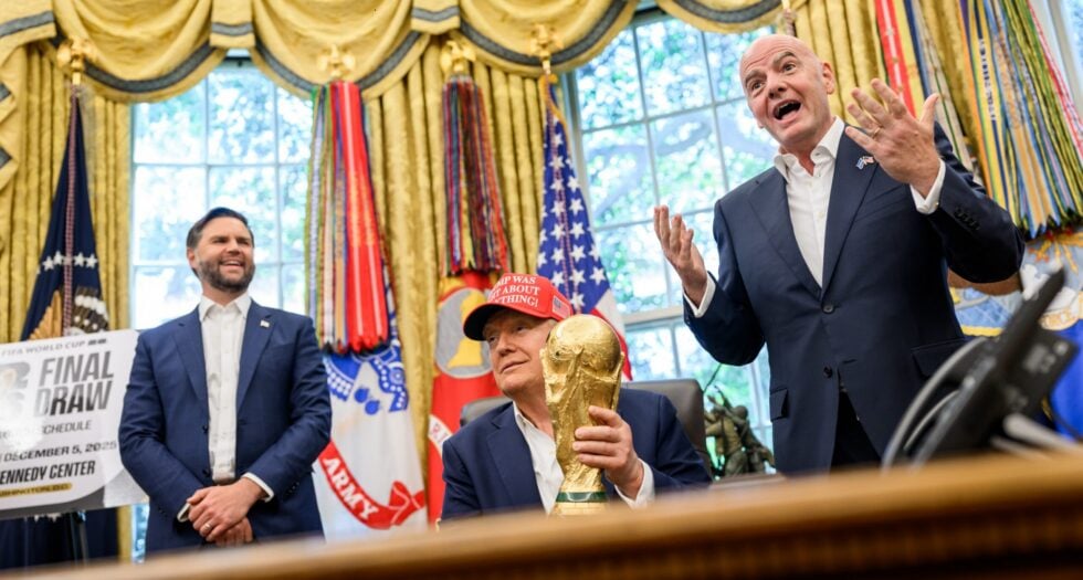 Three men in dark suits celebrate in a room with gold drapes and flags; one holds a large gold trophy while another gestures enthusiastically.