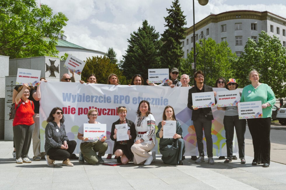 Group of diverse protesters holding placards and a large banner in a city square.