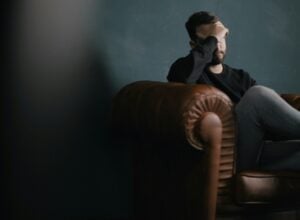 Man sitting on a brown leather couch, hand over his forehead, in a dim room, looking distressed.