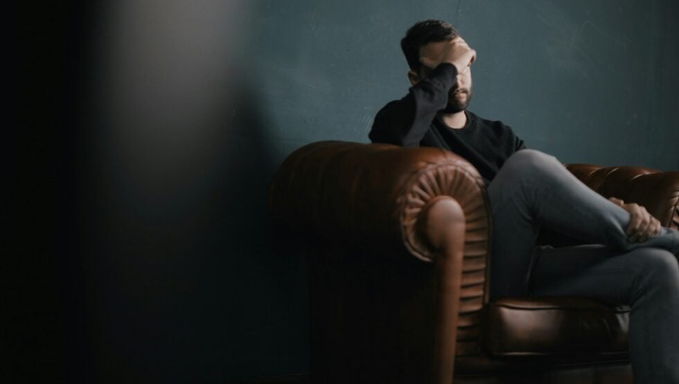 Man sitting on a brown leather couch, hand over his forehead, in a dim room, looking distressed.