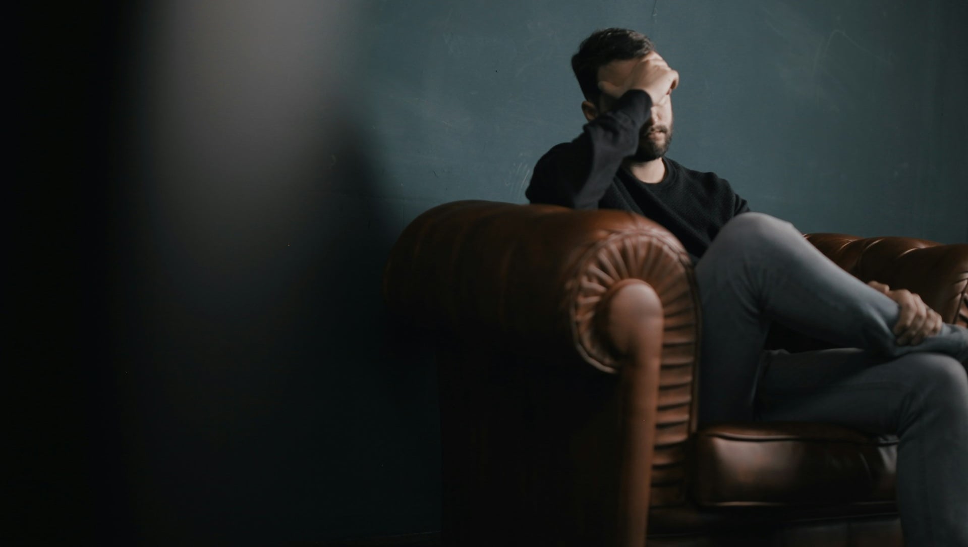 Man sitting on a brown leather couch, hand over his forehead, in a dim room, looking distressed.