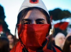 Close-up of a person wearing a red bandana over the mouth and a white cap at a protest.