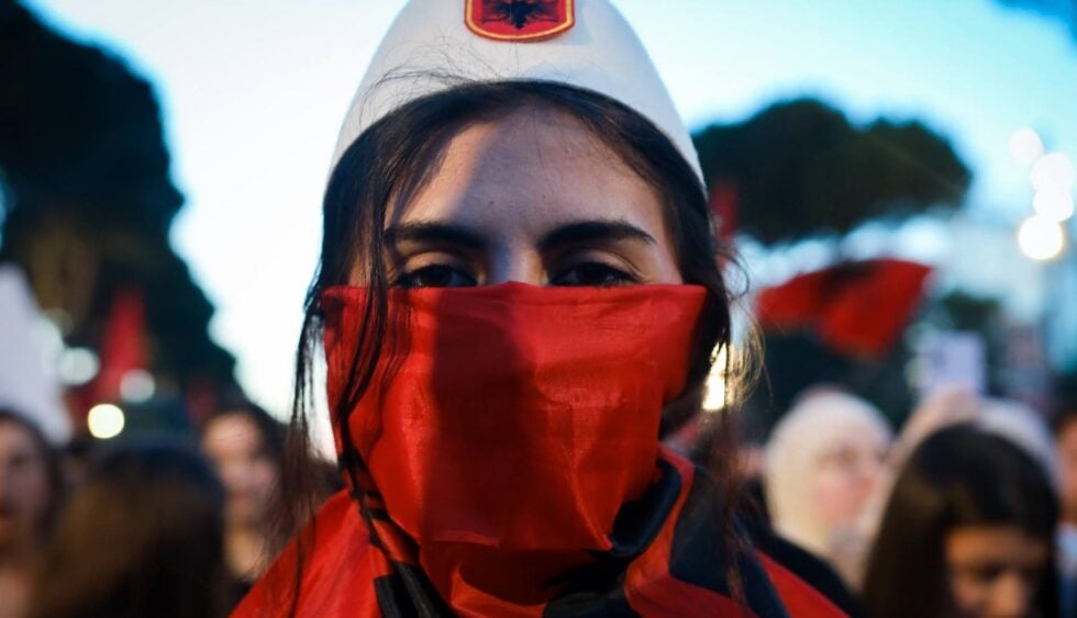 Close-up of a person wearing a red bandana over the mouth and a white cap at a protest.
