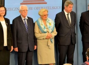 Five formally dressed individuals stand in a line on a stage, posing for a photo against a blue backdrop with red text.