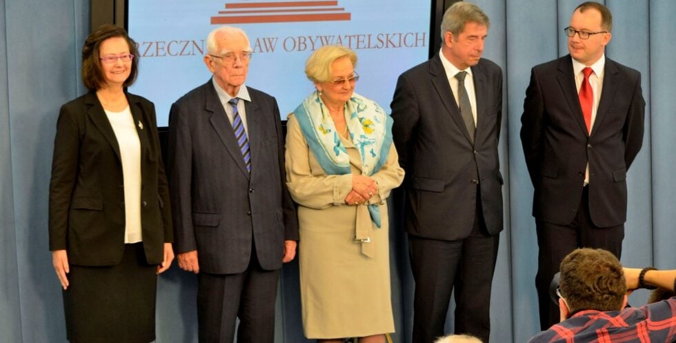 Five formally dressed individuals stand in a line on a stage, posing for a photo against a blue backdrop with red text.