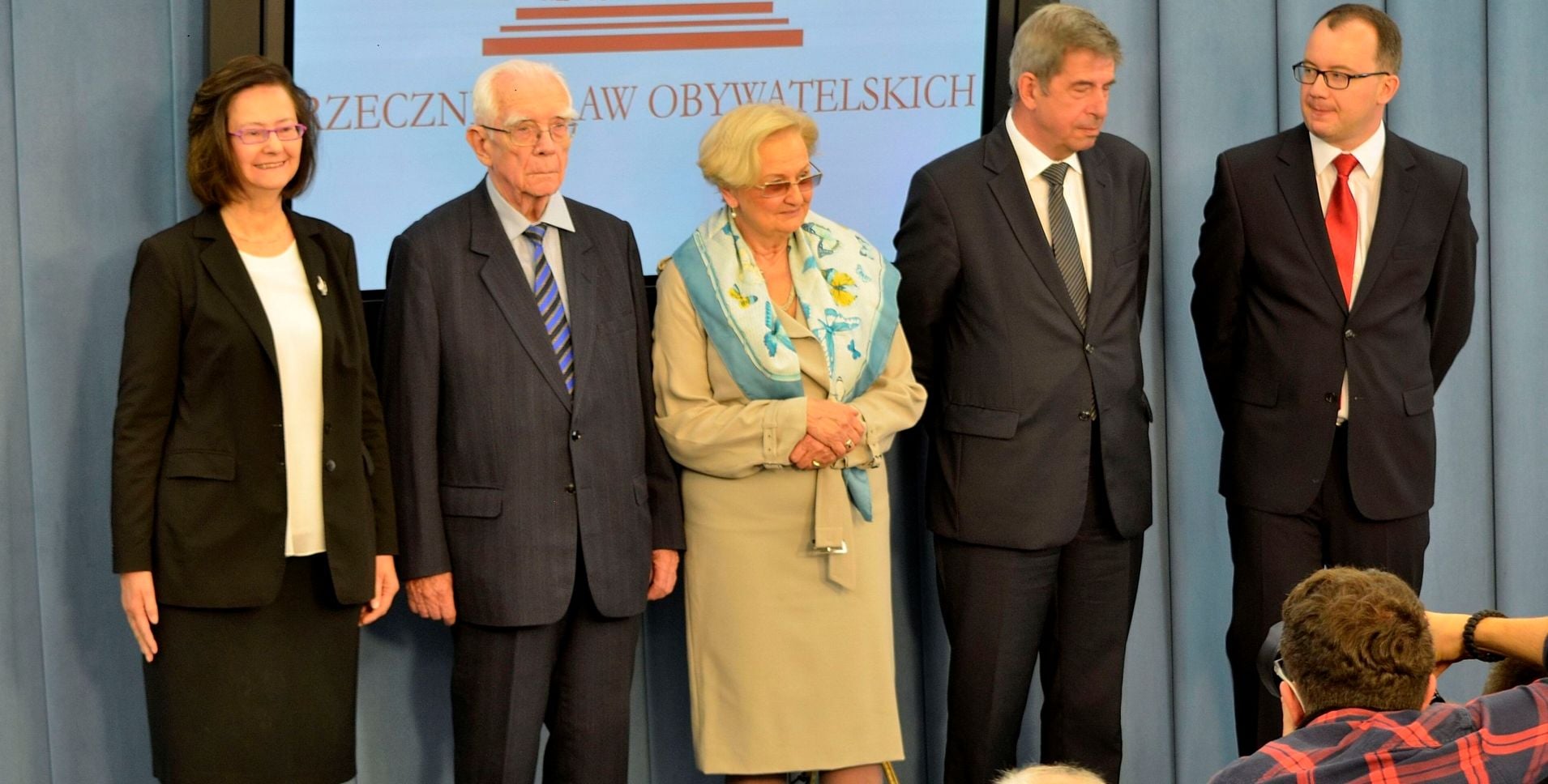 Five formally dressed individuals stand in a line on a stage, posing for a photo against a blue backdrop with red text.