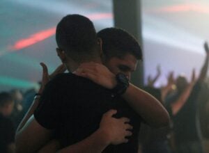 Two people embrace in a close hug at a dimly lit party with hands raised in the background.