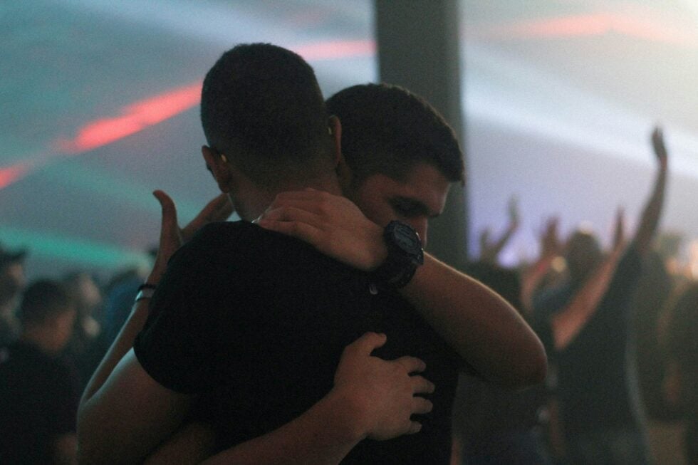 Two people embrace in a close hug at a dimly lit party with hands raised in the background.