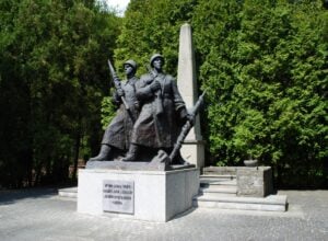 Bronze statue of two soldiers with rifles, standing in front of a tall obelisk in a paved square, surrounded by greenery.