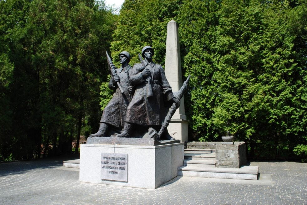 Bronze statue of two soldiers with rifles, standing in front of a tall obelisk in a paved square, surrounded by greenery.