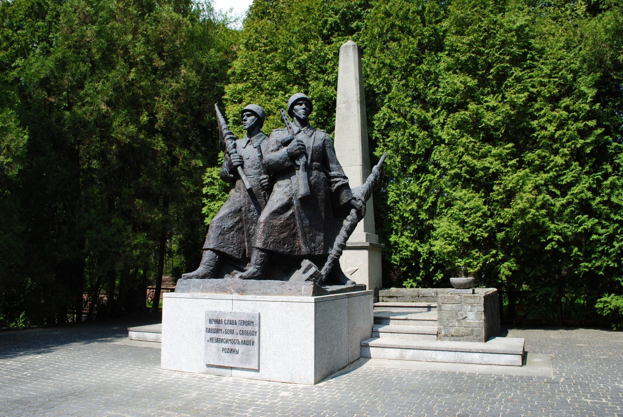 Bronze statue of two soldiers with rifles, standing in front of a tall obelisk in a paved square, surrounded by greenery.