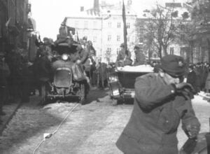 Black-and-white photo of a military parade on a city street, soldiers atop a vehicle, crowds lining the sidewalks in winter coats.
