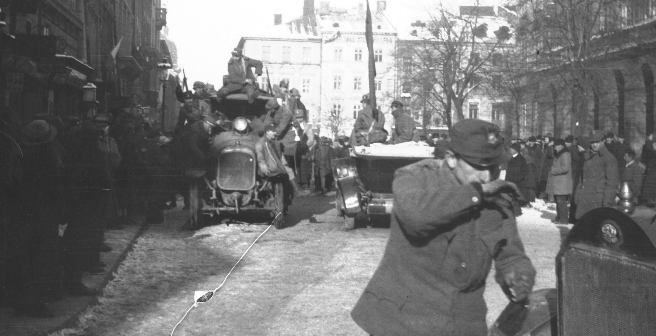 Black-and-white photo of a military parade on a city street, soldiers atop a vehicle, crowds lining the sidewalks in winter coats.