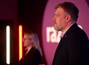 Profile of a man in a dark suit speaking at an event, pink and red stage lighting with vertical neon lights in the background.