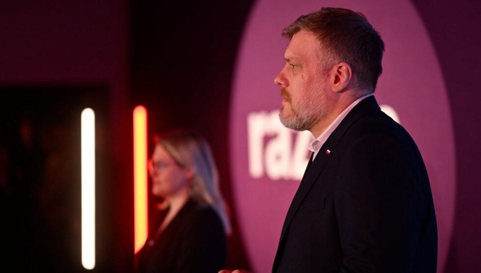 Profile of a man in a dark suit speaking at an event, pink and red stage lighting with vertical neon lights in the background.