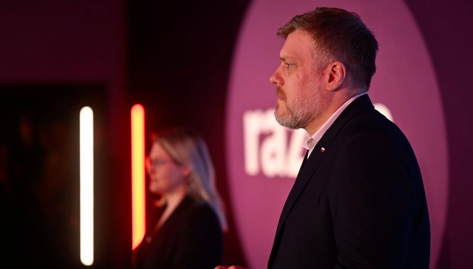 Profile of a man in a dark suit speaking at an event, pink and red stage lighting with vertical neon lights in the background.
