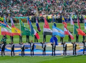 Flag bearers march with colorful national flags on a football field during a ceremony, with a packed stadium in the background.
