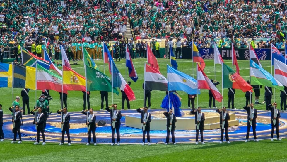 Flag bearers march with colorful national flags on a football field during a ceremony, with a packed stadium in the background.
