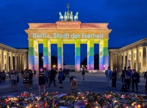 Rainbow-lit Brandenburg Gate at night with a crowd gathered and a floral memorial in the foreground.