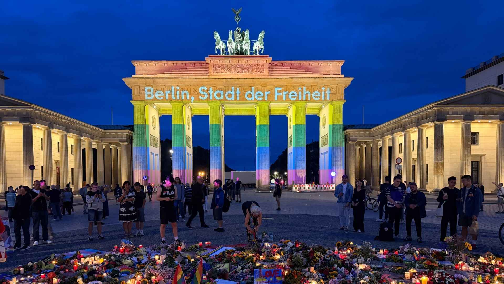 Rainbow-lit Brandenburg Gate at night with a crowd gathered and a floral memorial in the foreground.
